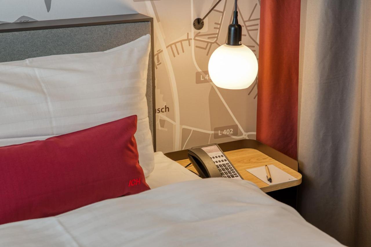 Intercityhotel Berlin Airport Ber Terminal 1 & 2 Schoenefeld Luaran gambar The photo shows a cozy bedroom setup. There's a neatly made bed with white bedding and a red decorative pillow. Beside the bed, a small bedside table holds a telephone and a notebook or notepad, along with a pen. A round pendant light hangs above the